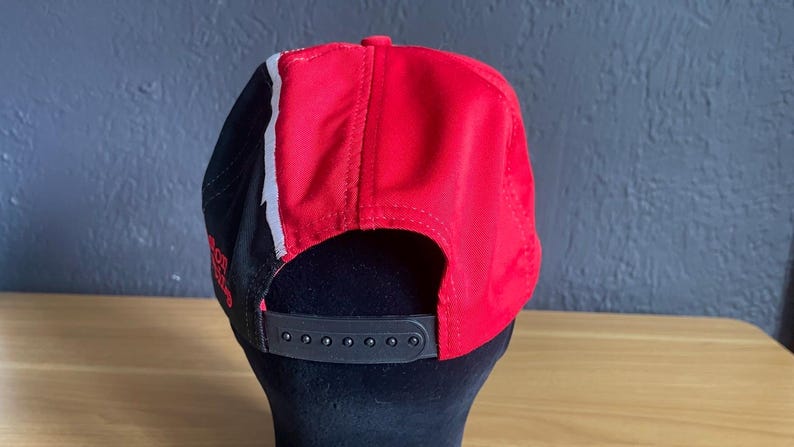 K&ouml;nnte beinhalten: Rote und schwarze Baseballkappe mit wei&szlig;em Streifen. Die Kappe hat einen schwarzen, verstellbaren Riemen und einen gebogenen Schirm. Die R&uuml;ckseite der Kappe ist rot, die Seiten sind schwarz mit einem wei&szlig;en Streifen.