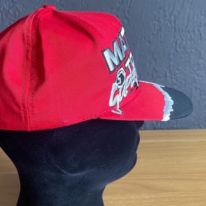 K&ouml;nnte beinhalten: Rote Baseballkappe mit schwarzem Schirm. Die Kappe hat wei&szlig;e und silberne Schrift und Grafiken auf der Vorderseite. Die Kappe wird auf einem schwarzen Schaufensterkopf pr&auml;sentiert.
