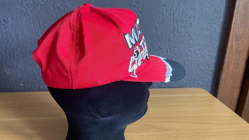 K&ouml;nnte beinhalten: Rote Baseballkappe mit schwarzem Schirm. Die Kappe hat wei&szlig;e und silberne Schrift und Grafiken auf der Vorderseite. Die Kappe wird auf einem schwarzen Schaufensterkopf pr&auml;sentiert.
