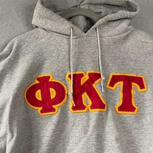 Pode incluir: Um moletom com capuz cinza mescla apresentando as letras gregas Phi Kappa Tau em tecido vermelho com uma borda amarela costurada no centro do peito. O moletom inclui um cordão cinza correspondente e um bolso frontal padrão.