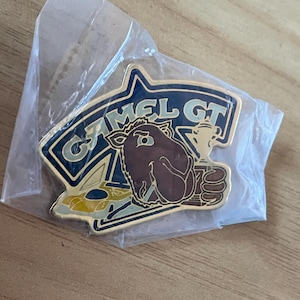 May include: A Camel GT pin in a clear plastic bag. The pin features a brown camel holding a trophy, with the words "CAMEL GT" in a blue and gold design. A yellow and blue race car is also visible.