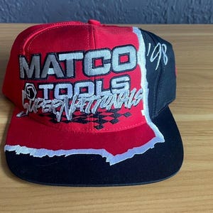 K&ouml;nnte beinhalten: Rote, schwarze und wei&szlig;e Baseballkappe mit dem Text "MATCO TOOLS SUPER NATIONALS" in Wei&szlig; und Silber. Die Kappe hat einen schwarzen Schirm und eine schwarze Seitenfl&auml;che mit der Zahl "'98" in Wei&szlig;.