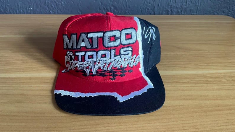 K&ouml;nnte beinhalten: Rote, schwarze und wei&szlig;e Baseballkappe mit dem Text "MATCO TOOLS SUPER NATIONALS" in Wei&szlig; und Silber. Die Kappe hat einen schwarzen Schirm und eine schwarze Seitenfl&auml;che mit der Zahl "'98" in Wei&szlig;.