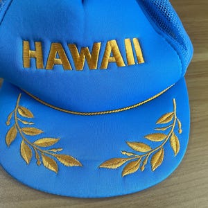 Puede incluir: Gorra de camionero azul con la palabra "HAWAII" bordada en dorado. La visera presenta bordados de hojas doradas y un ribete de cuerda dorada. La parte superior del sombrero es de malla azul.
