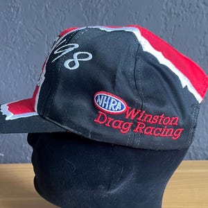 K&ouml;nnte beinhalten: Schwarze Baseballkappe mit roten und wei&szlig;en Akzenten. Sie tr&auml;gt den Schriftzug "48" in wei&szlig;er Schrift und das Logo "NHRA Winston Drag Racing" in Rot. Die Kappe wird auf einem schwarzen Schaufensterkopf pr&auml;sentiert.