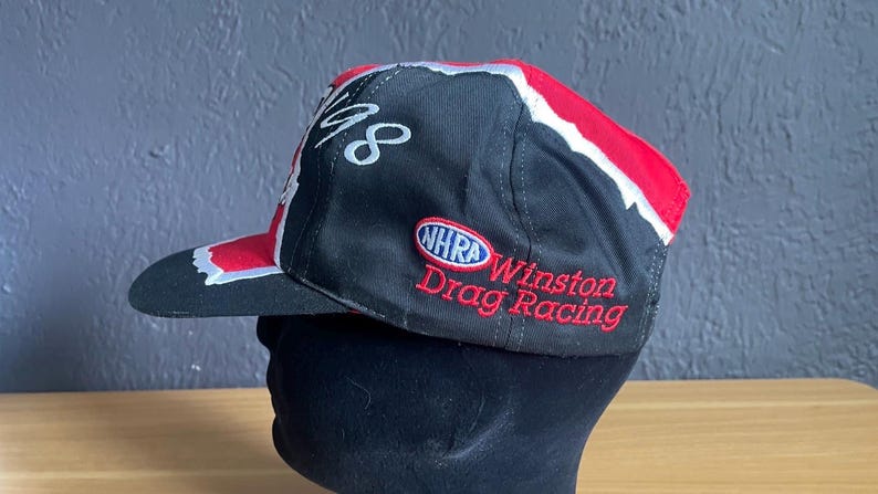 K&ouml;nnte beinhalten: Schwarze Baseballkappe mit roten und wei&szlig;en Akzenten. Sie tr&auml;gt den Schriftzug "48" in wei&szlig;er Schrift und das Logo "NHRA Winston Drag Racing" in Rot. Die Kappe wird auf einem schwarzen Schaufensterkopf pr&auml;sentiert.