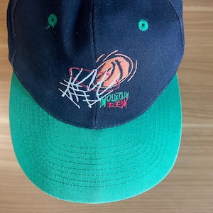 Puede incluir: Gorra de béisbol azul marino con visera verde. La gorra presenta un diseño bordado con el texto "MOUNTAIN DEW" en verde y rojo, junto con un gráfico de baloncesto. La gorra tiene un ajuste estructurado.