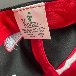 K&ouml;nnte beinhalten: Schwarze Baseballkappe mit rotem Schirm und Innenfutter. Das wei&szlig;e Etikett tr&auml;gt den Markennamen "Kudzu" und den Text "ONE SIZE FITS MOST MADE IN TAIWAN R.O.C. 65% POLYESTER 35% COTTON."
