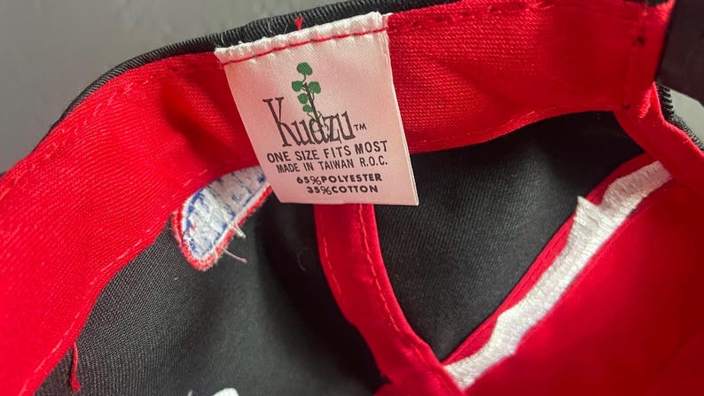 K&ouml;nnte beinhalten: Schwarze Baseballkappe mit rotem Schirm und Innenfutter. Das wei&szlig;e Etikett tr&auml;gt den Markennamen "Kudzu" und den Text "ONE SIZE FITS MOST MADE IN TAIWAN R.O.C. 65% POLYESTER 35% COTTON."