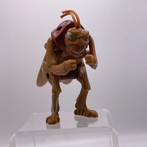 Puede incluir: Una figura de acción de plástico marrón de un personaje similar a un saltamontes con un caparazón marrón oscuro, puños cerrados y dos antenas de cordón naranja en la parte superior de la cabeza. La figura tiene una expresión facial severa.
