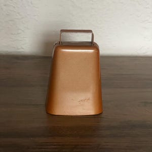 May include: A bronze-colored cowbell with a rectangular shape and a handle at the top. The bell has a smooth, metallic finish and sits on a dark wooden surface. The background is a textured, off-white wall.