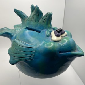 May include: A blue and green ceramic fish-shaped coin bank with a coin slot on the top. The fish features raised fins, a pointed tail, and large, protruding eyes with white and purple accents. The surface has a smooth, glossy finish.