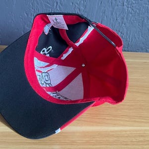 K&ouml;nnte beinhalten: Rote und schwarze Baseballkappe mit gebogenem Schirm. Die Innenseite ist rot und wei&szlig; mit schwarzen Akzenten. Die Kappe liegt auf einer Holzoberfl&auml;che.