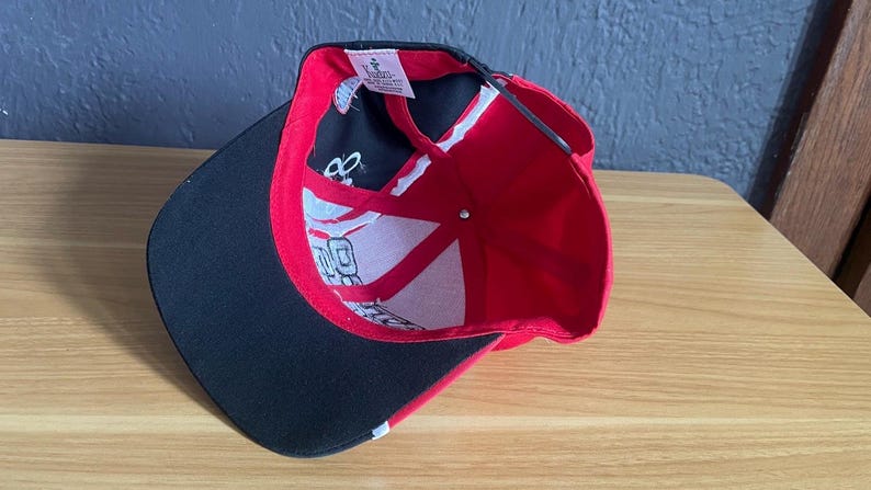 K&ouml;nnte beinhalten: Rote und schwarze Baseballkappe mit gebogenem Schirm. Die Innenseite ist rot und wei&szlig; mit schwarzen Akzenten. Die Kappe liegt auf einer Holzoberfl&auml;che.