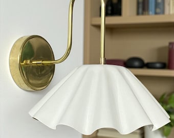 May include: A wall-mounted lamp featuring a polished brass arm that curves upward and outward from a circular brass base. The lamp includes a wide, white, pleated shade with a scalloped edge that directs light downward from the fixture.