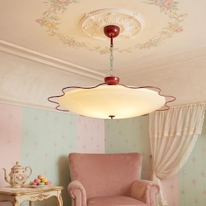 May include: A vintage-style ceiling light fixture with a cream-colored, scalloped shade and a dark red trim. The fixture hangs from a chain attached to a decorative ceiling medallion. The room features pink and green walls, a pink armchair, and floral accents.
