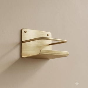 Op de afbeelding: Een wandhouder van geborsteld messing met een rechthoekige achterplaat met twee zichtbare schroefgaten aan de bovenkant, een U-vormige steunbeugel en een platte, licht gebogen onderste plank om voorwerpen stevig op hun plaats te houden.