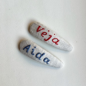 May include: Two fabric hair clips with embroidered names. One clip has the name "Veja" in red thread, and the other has "Aida" in blue thread. The clips are light grey and sit on a white surface.