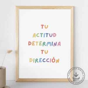 Puede incluir: Una impresión enmarcada con fondo blanco y texto en colores arcoíris que dice "Tu Actitud Determina Tu Dirección". El texto está en una fuente redonda y divertida. El marco es de madera clara.