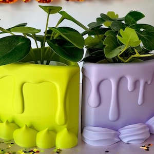 May include: Two ceramic planters with a drip glaze design. The planter on the left is yellow and the planter on the right is purple. Both planters have a decorative ruffle at the bottom.