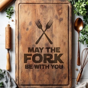 Op de afbeelding: Een rechthoekige houten snijplank met een donkerbruine nerf, voorzien van een gegraveerd ontwerp van twee gekruiste vorken en de tekst "MAY THE FORK BE WITH YOU" in het midden. De plank heeft een gegroefde rand langs de zijkanten.