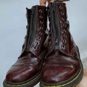 May include: A pair of burgundy leather boots with black zipper detailing and laces. The boots have a yellow stitch around the sole.