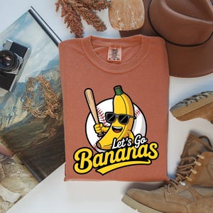 May include: A rust-colored t-shirt featuring a cartoon banana wearing sunglasses and holding a baseball bat. The shirt has the text "Let's Go Bananas" in a yellow and black font. The image also includes a brown hat, boots, and a camera.