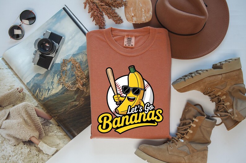 May include: A rust-colored t-shirt featuring a cartoon banana wearing sunglasses and holding a baseball bat. The shirt has the text "Let's Go Bananas" in a yellow and black font. The image also includes a brown hat, boots, and a camera.