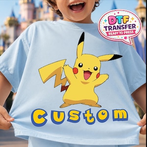 May include: Light blue t-shirt featuring a yellow Pikachu graphic with red cheeks and black-tipped ears. The word "CUSTOM" is printed in yellow with a blue outline. A "DTF TRANSFER READY TO PRESS" graphic is in the upper right corner.