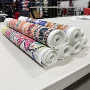 May include: Several rolls of printed fabric are stacked on a white table. The fabric rolls feature various designs, including sports team logos and cartoon graphics. The rolls are white with colorful printed designs.