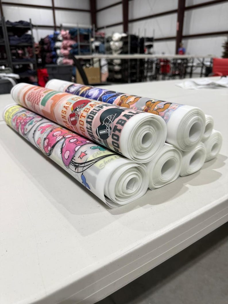 May include: Several rolls of printed fabric are stacked on a white table. The fabric rolls feature various designs, including sports team logos and cartoon graphics. The rolls are white with colorful printed designs.