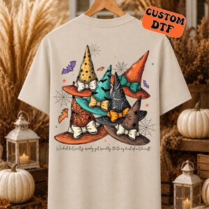 May include: A beige short-sleeve t-shirt featuring a graphic print of five colorful witch hats with bows, spiderwebs, and bats. The text below the hats reads, "Wicked but pretty, spooky yet sparkly, that's my kind of witchcraft."