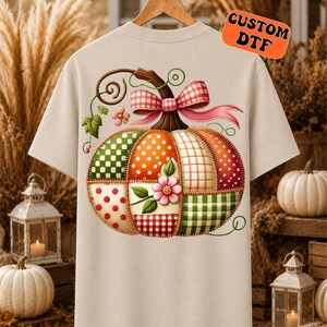 May include: A beige short-sleeved t-shirt featuring a large graphic of a patchwork pumpkin on the back. The pumpkin is composed of various patterned squares in orange, green, and white, accented with a pink gingham bow and a small pink flower.