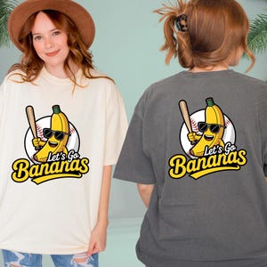 May include: Cream-colored and charcoal gray t-shirts featuring a cartoon banana wearing sunglasses and holding a baseball bat. The text "Let's Go Bananas" is below the banana graphic. The shirts are casual and unisex.