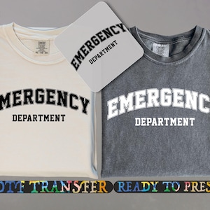 May include: Two t-shirts, one cream and one gray, each with the words "EMERGENCY DEPARTMENT" in a bold, arched font. A matching design is on a small, square transfer sheet. The shirts are folded, and the design is centered.