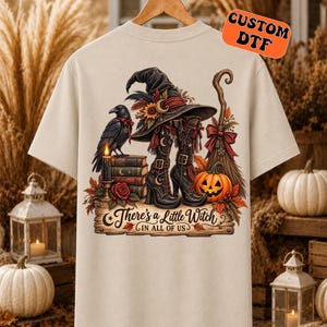 May include: A beige t-shirt featuring a graphic print of a black witch hat, black boots, a raven on a stack of books, a broom, and a jack-o'-lantern. The text reads, There's a Little Witch IN ALL OF US. The design includes autumn leaves and a rose.