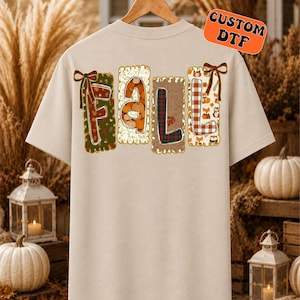 May include: A beige t-shirt featuring the word FALL in large, decorative letters. Each letter is contained within a rectangular border with distinct patterns including pumpkins, plaid, and woodland animals, accented by brown bows.