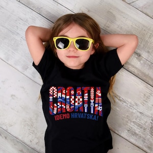 May include: A young person wearing a black t-shirt with the word "CROATIA" in a colorful, patterned design. The shirt also has the text "IDEMO HRVATSKA!" The person is wearing yellow sunglasses.