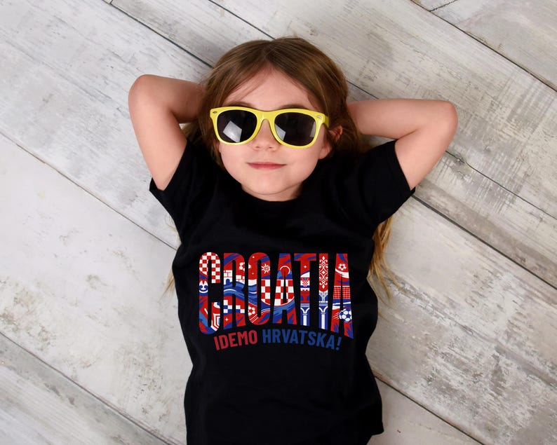 May include: A young person wearing a black t-shirt with the word "CROATIA" in a colorful, patterned design. The shirt also has the text "IDEMO HRVATSKA!" The person is wearing yellow sunglasses.