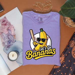 May include: A lavender t-shirt with a cartoon banana wearing sunglasses and holding a baseball bat. The text "Let's Go Bananas" is in yellow and black. The shirt is folded on a brown surface.