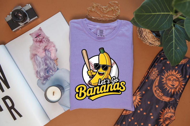 May include: A lavender t-shirt with a cartoon banana wearing sunglasses and holding a baseball bat. The text "Let's Go Bananas" is in yellow and black. The shirt is folded on a brown surface.
