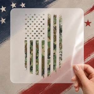 May include: A clear, rectangular stencil featuring a distressed American flag design. The flag's stripes are camouflage, and the stars are a mix of green and brown. The background has brushstrokes of red, white, and blue.