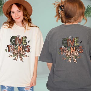 May include: Cream-colored and charcoal gray t-shirts with a graphic that says "GIVE THANKS". The graphic includes a bow, flowers, and fall colors. The shirts are oversized and casual.