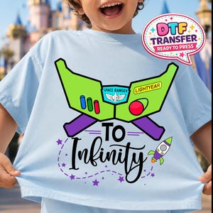 May include: Light blue t-shirt featuring a green and purple graphic of Buzz Lightyear's space ranger helmet, with the words "TO Infinity" and a rocket ship. The shirt also has the text "DTF TRANSFER READY TO PRESS".