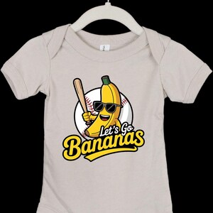 May include: A beige baby onesie with a cartoon banana wearing sunglasses and holding a baseball bat. The design includes a baseball and the text "Let's Go Bananas" in yellow and black.