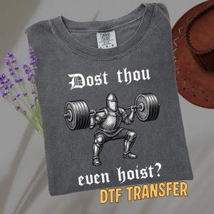 May include: Dark gray t-shirt with a graphic of a knight lifting weights, with the text "Dost thou even hoist?" and "DTF TRANSFER". The shirt has a crew neck and a Comfort Colors tag.