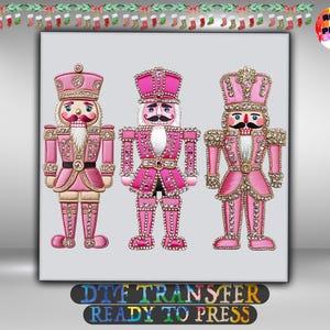 May include: Three nutcracker figures in shades of pink, each wearing a tall hat and uniform adorned with sparkling rhinestone accents. The figures feature white beards, black mustaches, and gold-toned trim. The text reads DTF TRANSFER READY TO PRESS.