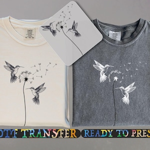 May include: Two t-shirts, one cream and one charcoal, each with a black and white hummingbird and dandelion design. The image includes a DTF transfer and the text "READY TO PRESS".