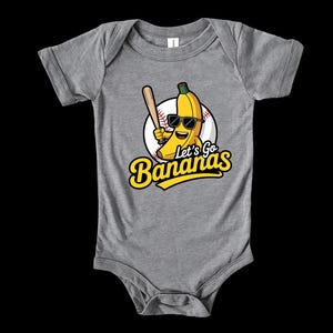 May include: A gray baby bodysuit with a graphic of a smiling banana wearing sunglasses and holding a baseball bat. The text "Let's Go Bananas" is in yellow script. The banana is set against a baseball.
