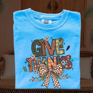 May include: A light blue Comfort Colors t-shirt with the words "GIVE THANKS" in a fall-themed design. The design includes a brown and green plaid bow, leaves, and floral accents. The shirt is a crew neck.